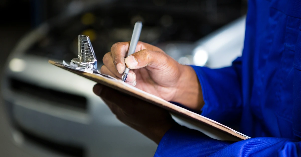 Mechanic in blue uniform writing on clipboard during car inspection in garage, checking vehicle details and estimating logbook service cost for maintenance