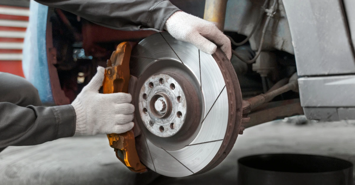 Gloved hands holding old brake pads next to a slotted rotor on a workshop floor — brake pad replacement on a lifted vehicle.